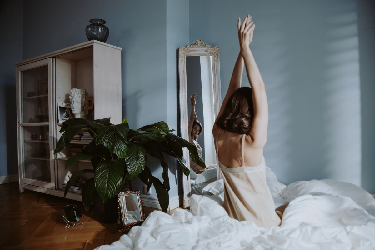 A woman in sleepwear stretches in a calm bedroom setting, reflected in a mirror