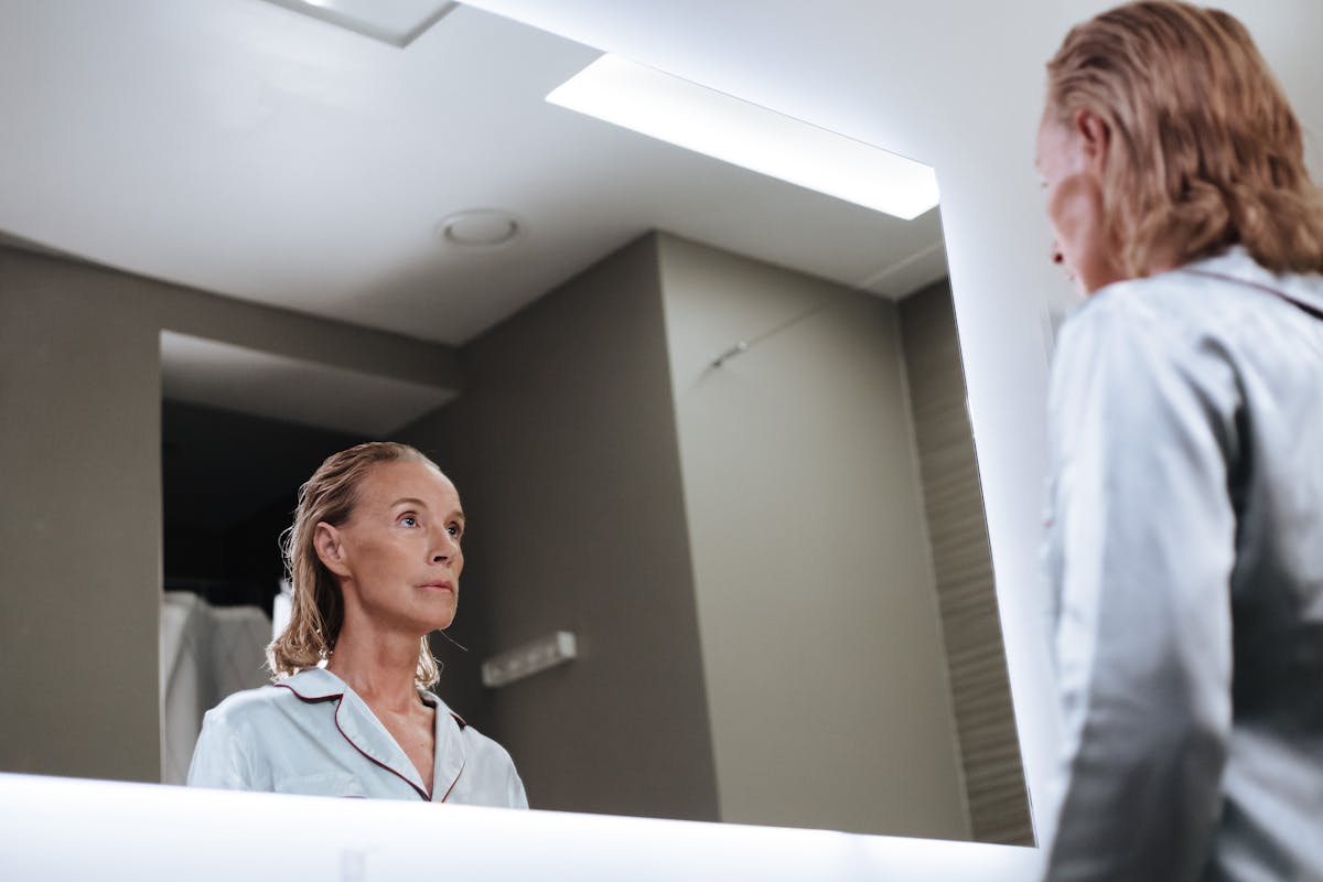 Woman looking introspectively at her bathroom mirror evoking the self-confrontational act of talking to a mirror and its psychological meaning A senior woman looks introspectively at herself in a modern bathroom mirror in quiet indoor lighting evoking the reflective quality of talking to a mirror and what it means