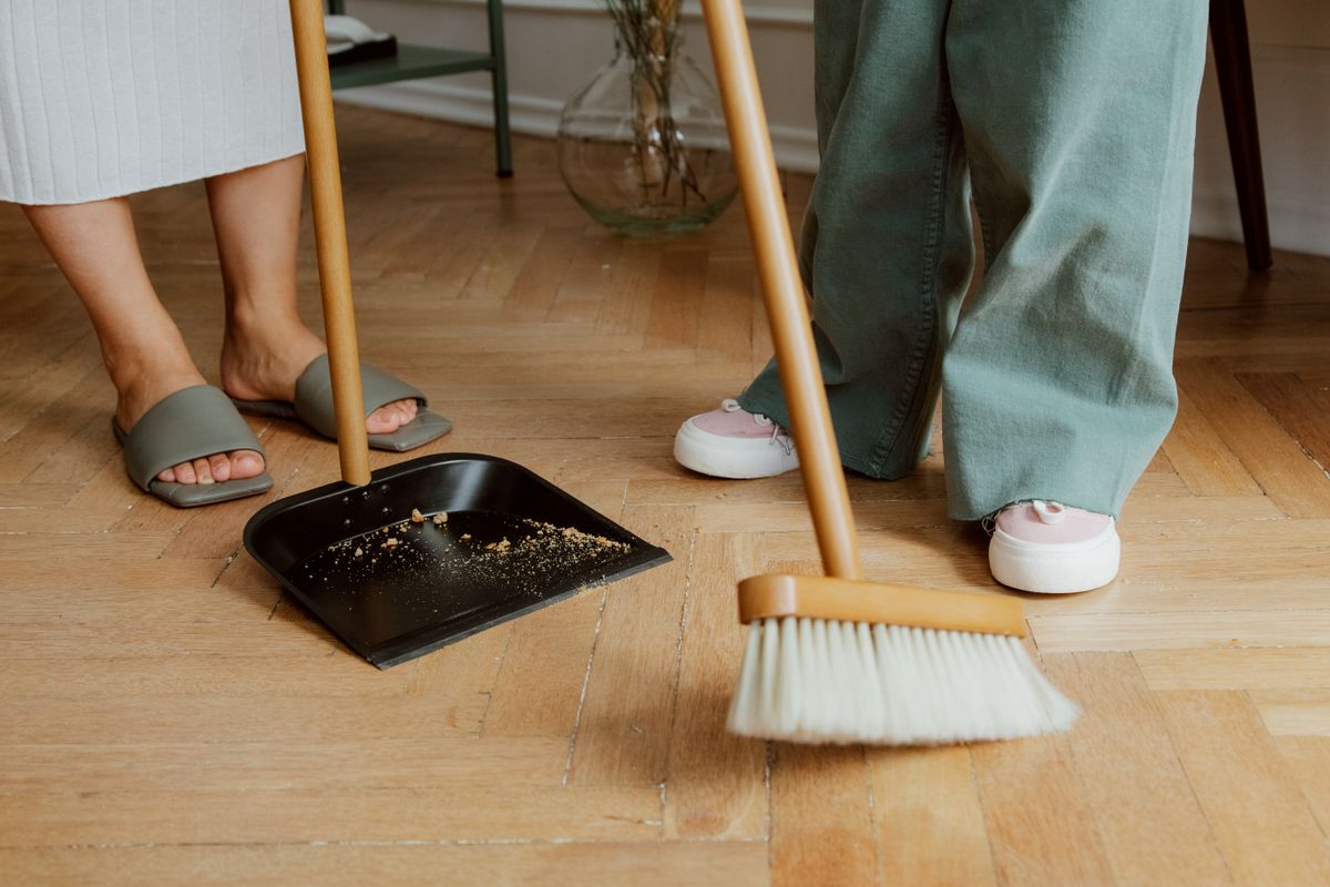 Close-up of people cleaning a wooden floor with broom and dustpan collecting broken glass representing the practical first steps in the broken mirror spiritual meaning