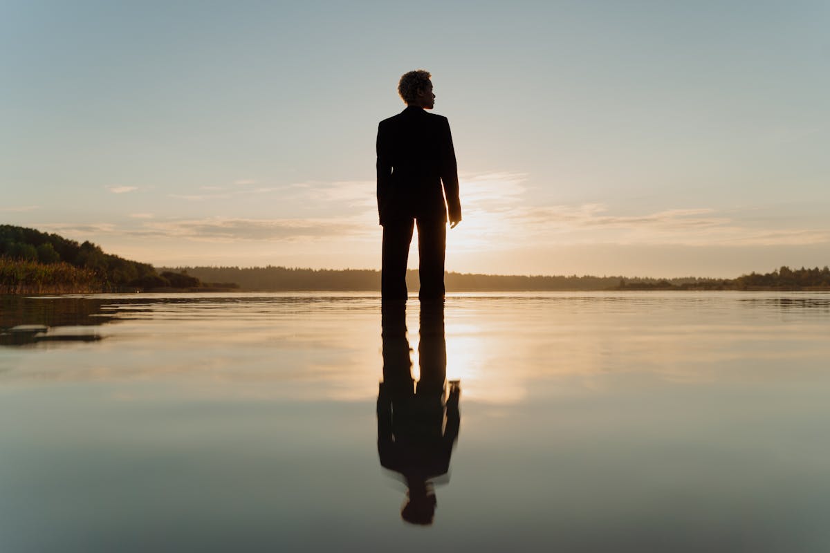 A dramatic silhouette of a figure standing on calm water at sunset reflected in the surface representing the shadow self and unconscious aspects revealed in mirror-in-water dreams
