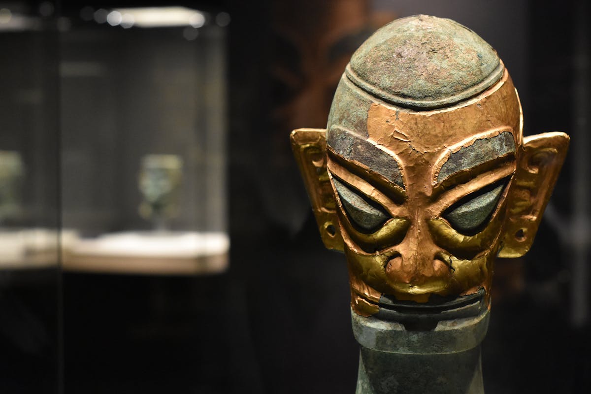 Close-up of an ancient bronze mask in a dimly lit museum display case representing the historical reality of ancient bronze mirrors that informed biblical mirror metaphors