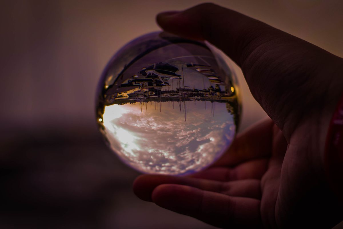 Hand holding a clear glass sphere reflecting a sunset harbor scene