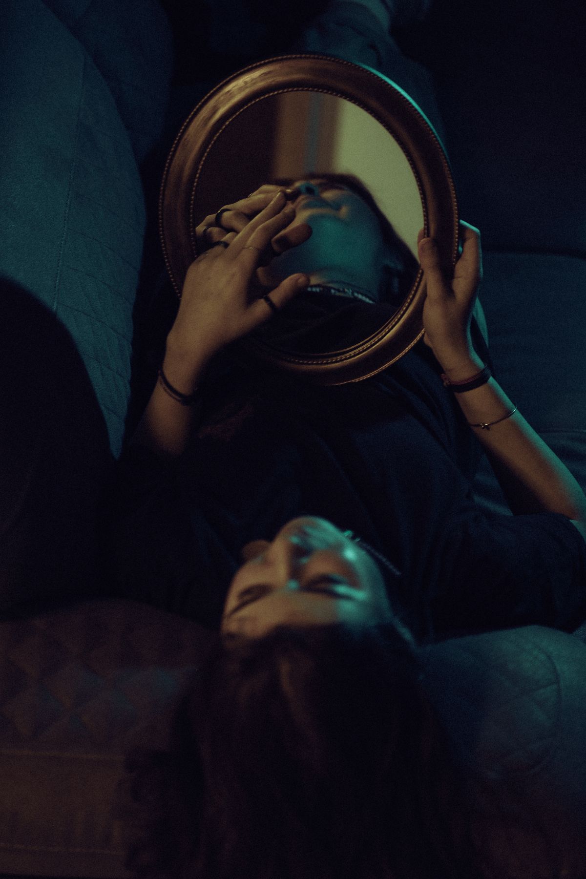 Moody interior scene with a reclining figure holding an oval handheld mirror reflecting dim window light