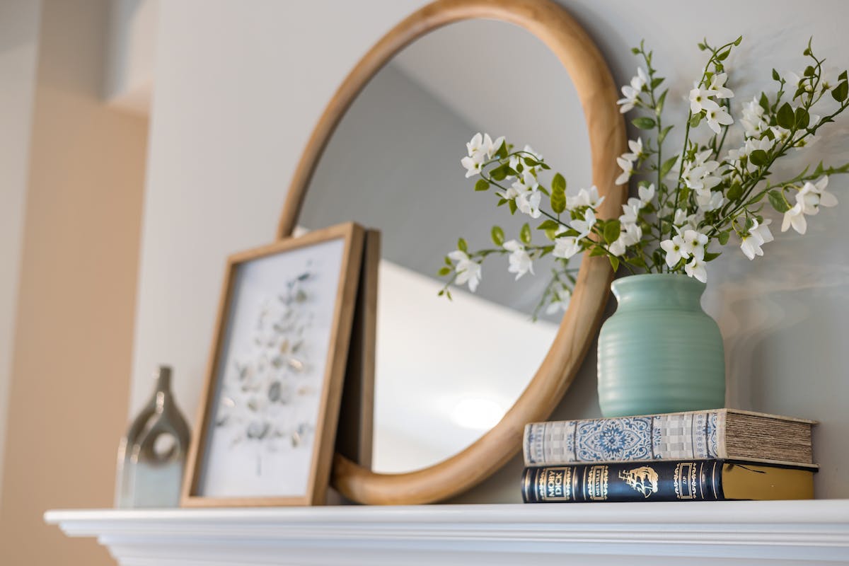 Framed decorative mirror styled with flowers and books illustrating round mirror frame styles Stylish interior with a framed mirror, vase of flowers, and books on a shelf, illustrating round mirror frame styles