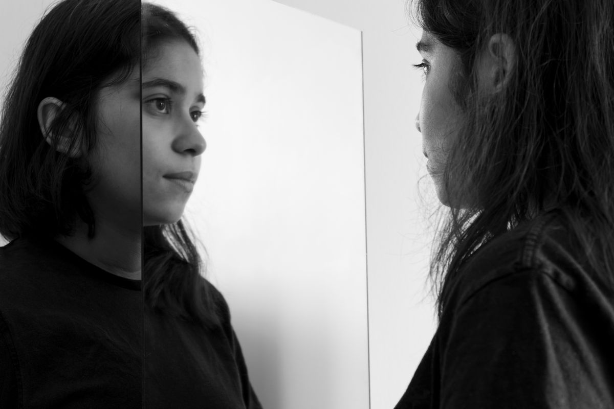 Young woman studying her reflection in a mirror in black and white Black and white photograph of a young woman reflecting on herself in a mirror indoors symbolising introspection and mirror dream psychology