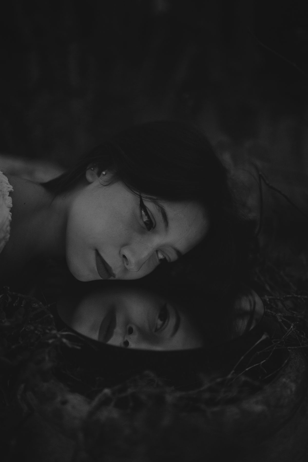 Black-and-white artistic portrait showing a reflective mirror surface doubling facial contours in subdued outdoor framing