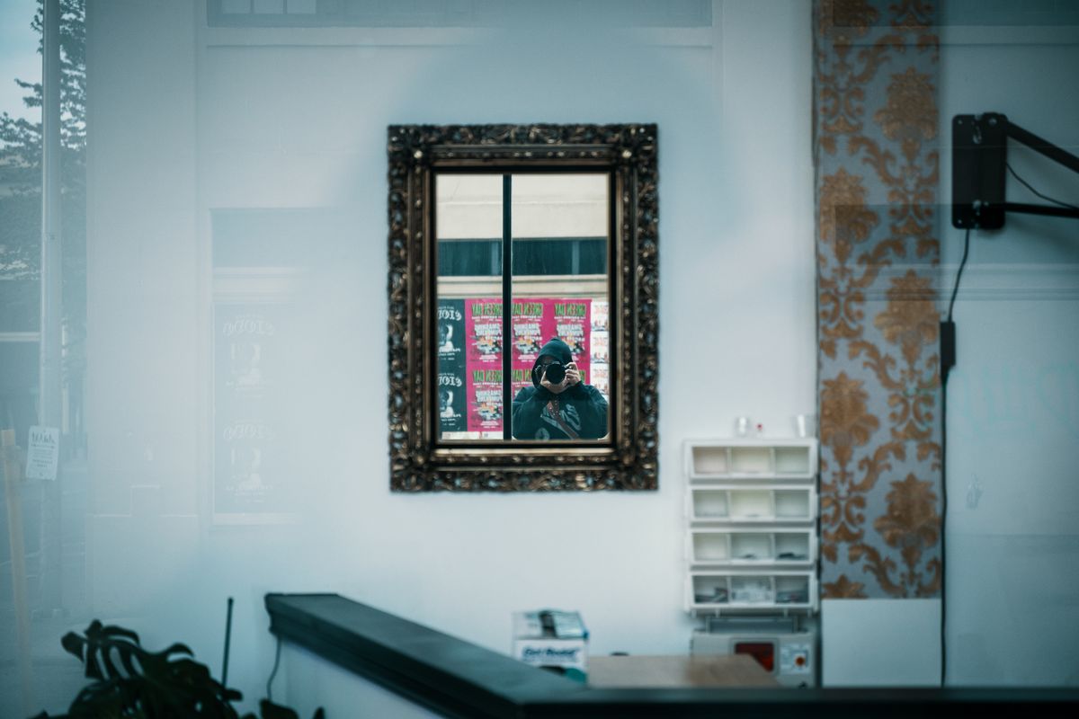 Person in dark knit sweater seen from side gazing into wall mirror in dim interior moody self reflection scene