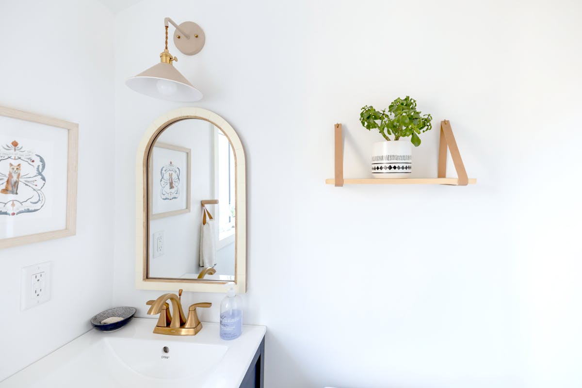 Gold-framed mirror in a bright bathroom illustrating arched mirror frame styles Bright bathroom interior with a gold-framed mirror and a decorative shelf, illustrating arched mirror frame styles