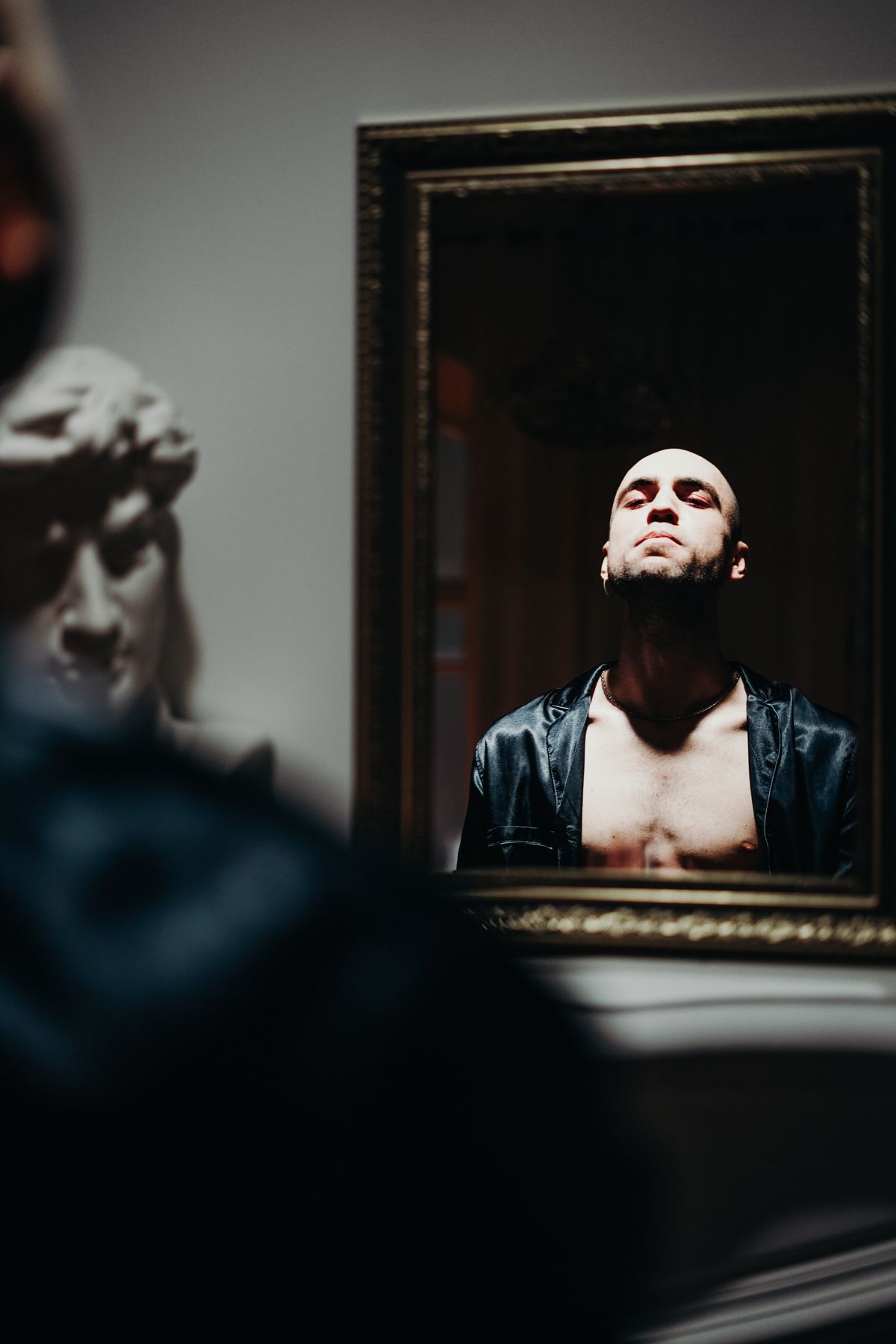 Bald person facing mirror reflection in near-dark room highlighting contemplative introspective atmosphere