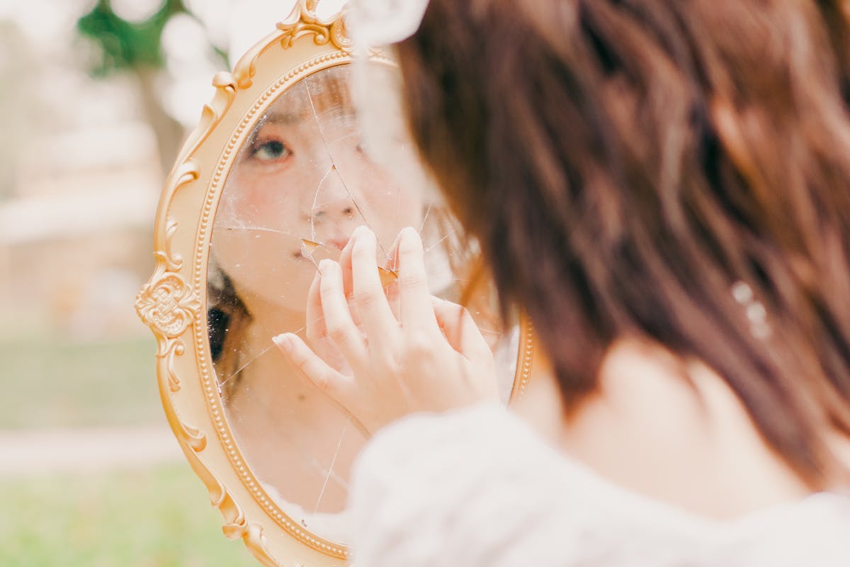 Young woman introspectively touching a broken mirror outdoors representing the broken mirror spiritual meaning of fractured self-image and identity