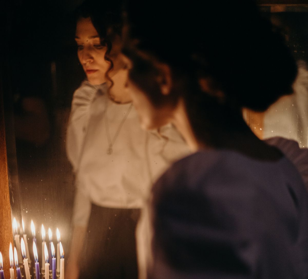 Woman in candlelit room gazing at mirror reflection conveying unease layered with folklore about doubled mirrors