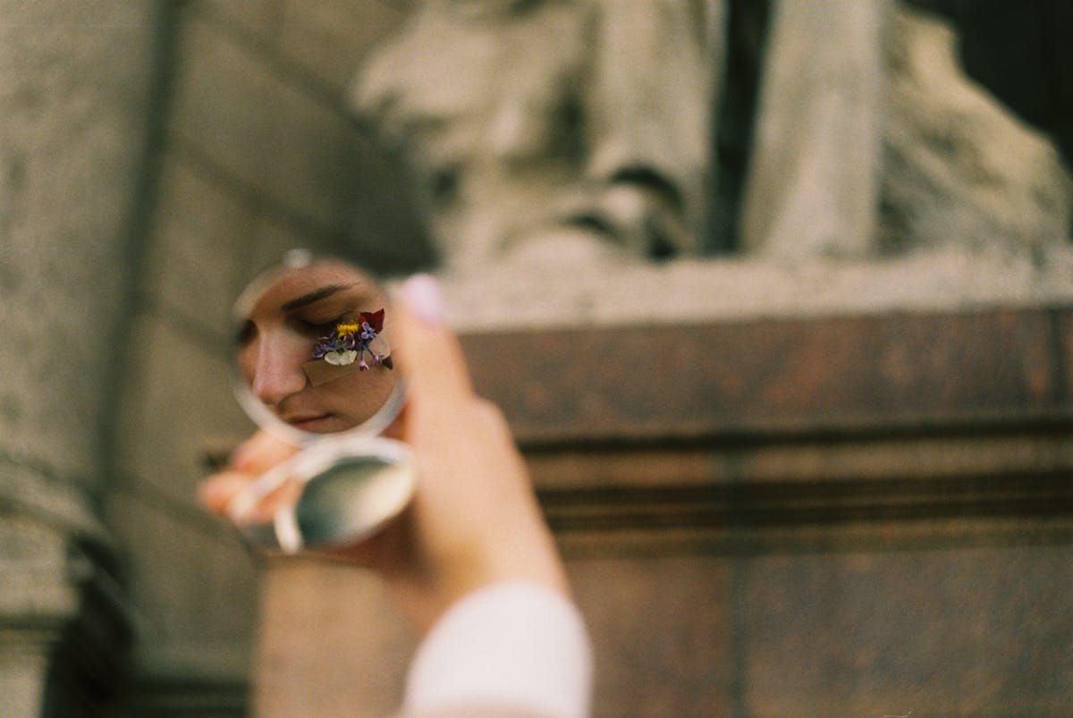 Face reflected in round mirror held by hand for spiritual mirror ritual and intention symbolism A person's face with flowers reflected in a round mirror held by a hand suggesting spiritual mirror ritual and intentional reflection
