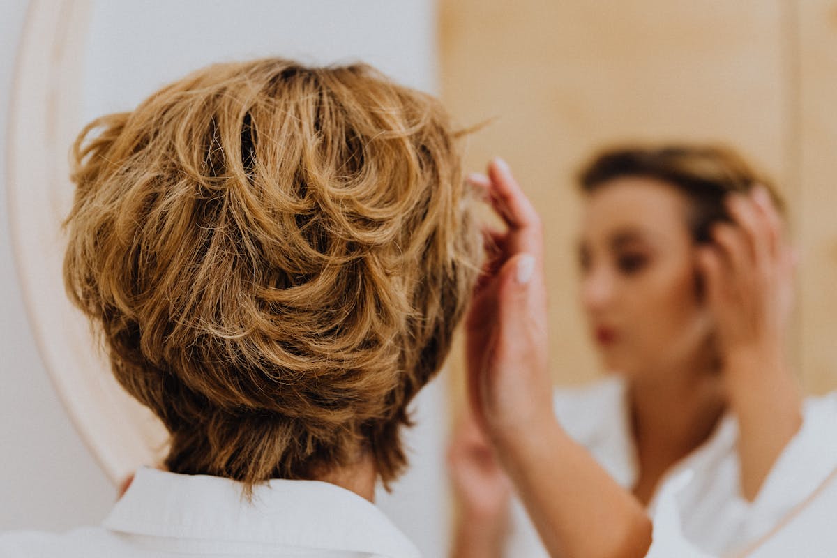 Back view of a woman styling her short hair while looking at her own reflection, representing everyday self-assessment at the mirror