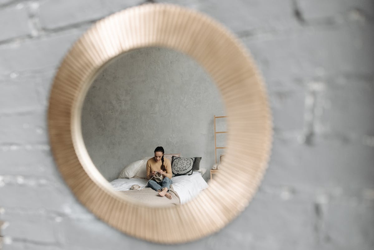 A woman knitting while sitting on a bed, reflected in a round wall mirror. A woman knitting while sitting on a bed, reflected in a round wall mirror.