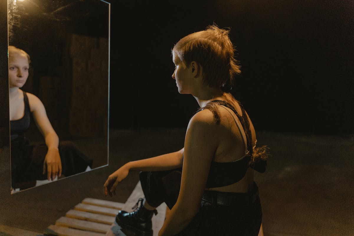 Woman with a shaved head sitting in front of a mirror in a dimly lit room reflecting on her image