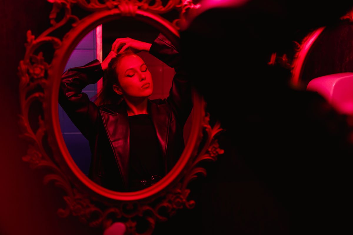 Figure adjusting appearance before moody red-lit mirror illustrating embodied portal metaphors and intentional gaze