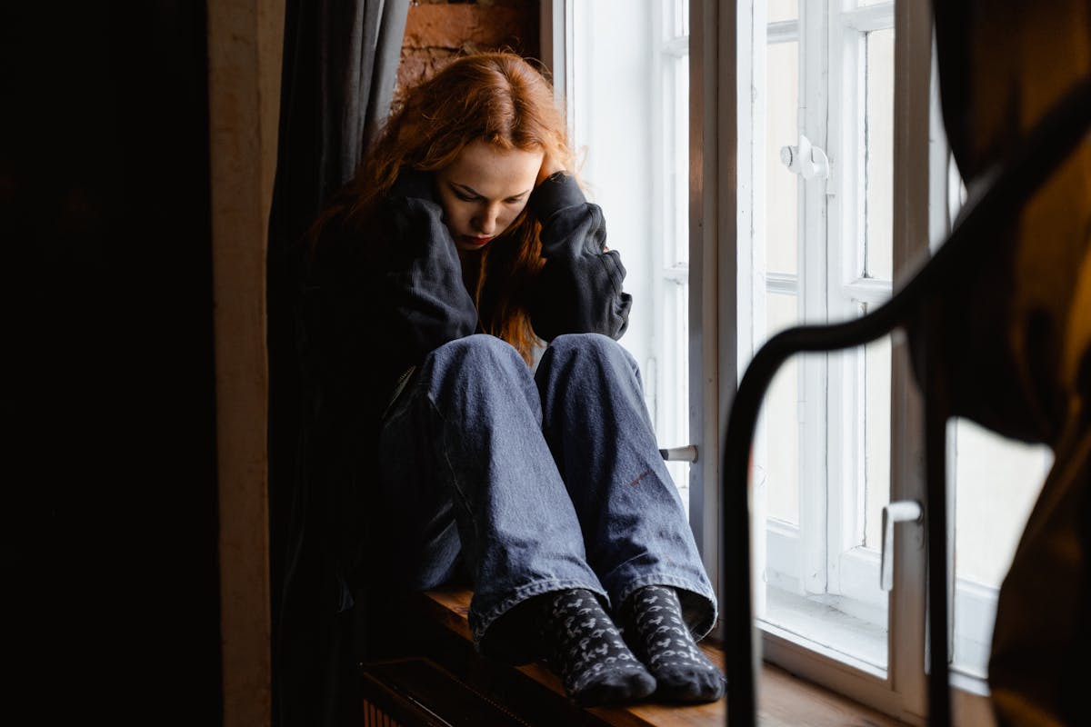 Pensive woman by window for mirror anxiety self-image and mental health discussion Woman sitting pensively by window representing mirror anxiety and the emotional impact of mirrors on mental health