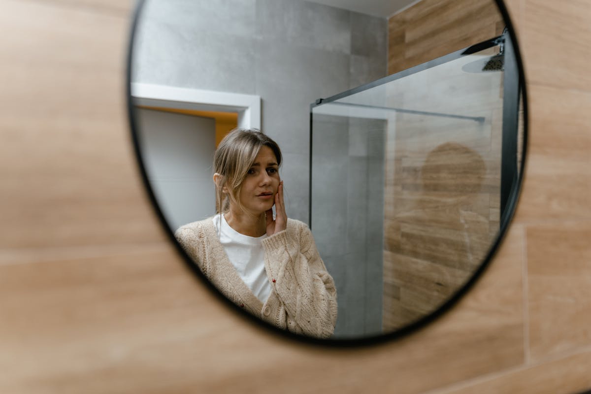 Woman in a neutral sweater looking at herself in a bathroom mirror in an introspective mood illustrating the fear of mirrors