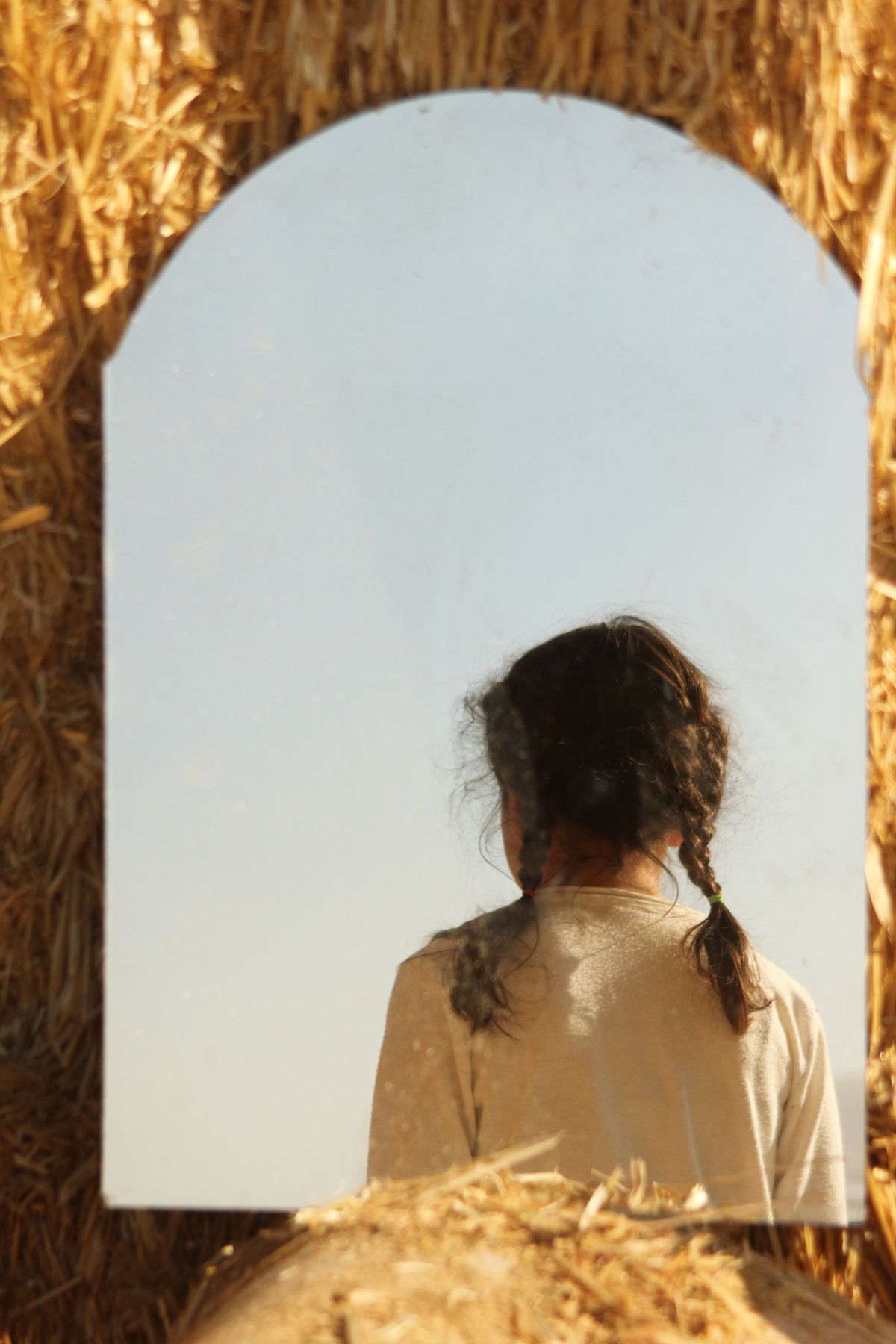 Girl with braided hair seen from behind reflected in mirror in soft outdoor hay field