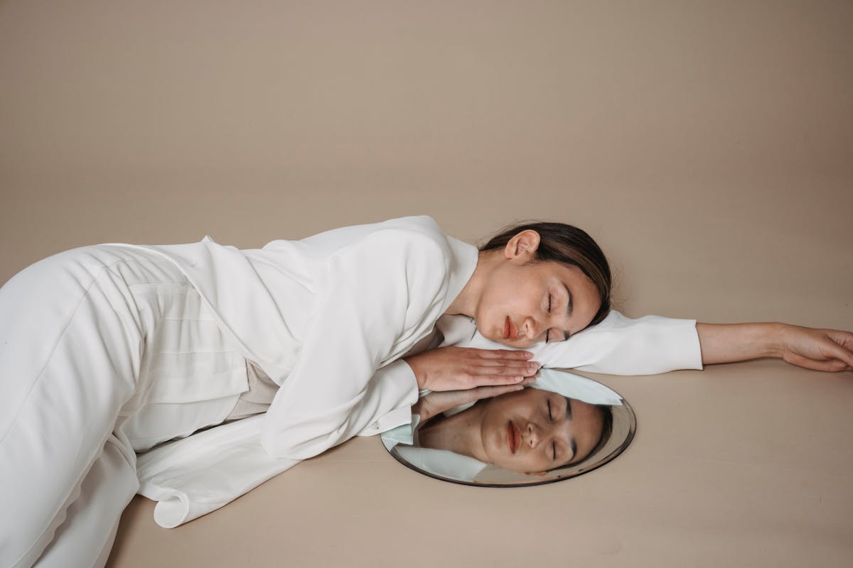 Artistic portrait of a woman in white suit lying on a mirror with eyes closed, evoking introspection and the deep symbolism of mirrors as reflections of the inner self