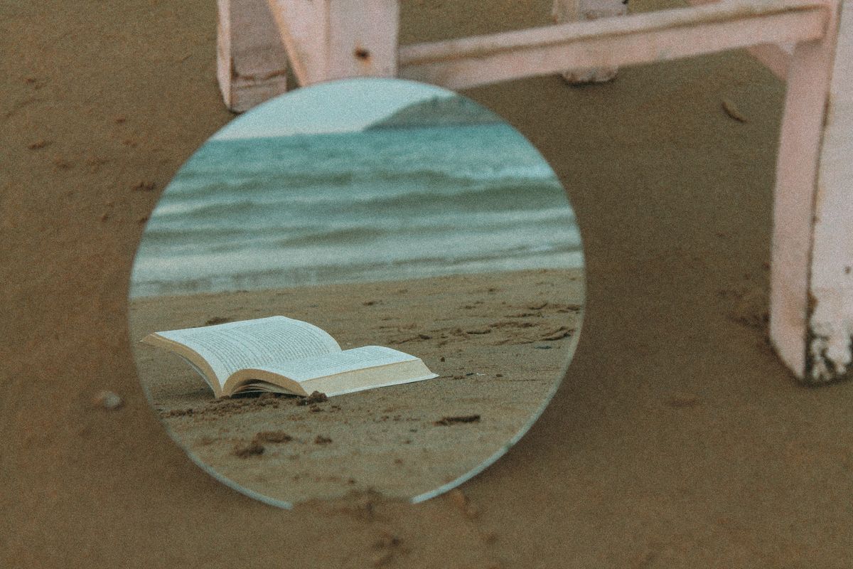 Round mirror reflecting an open book on a sandy beach showing how smooth reflective surfaces create coherent images through the law of reflection