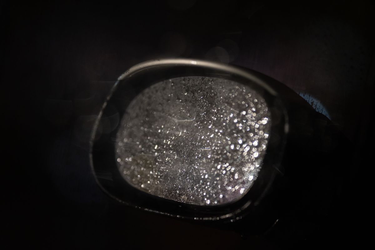 Rain-beaded automotive mirror glass macro emphasizing reflective boundary tension between viewer and wet darkness